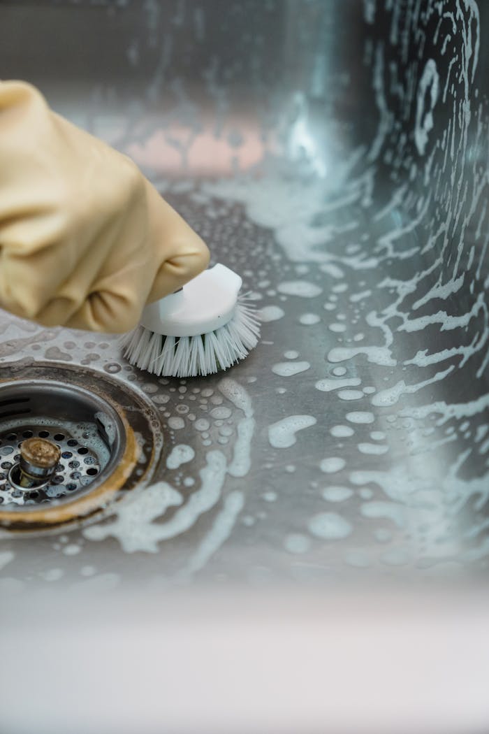 who-we-are Close-up of a hand in gloves scrubbing a soapy kitchen sink with a brush.