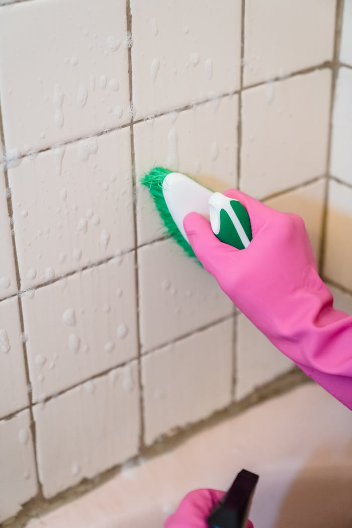 services-04 A hand in pink gloves scrubs bathroom tiles with a green brush to remove grime.